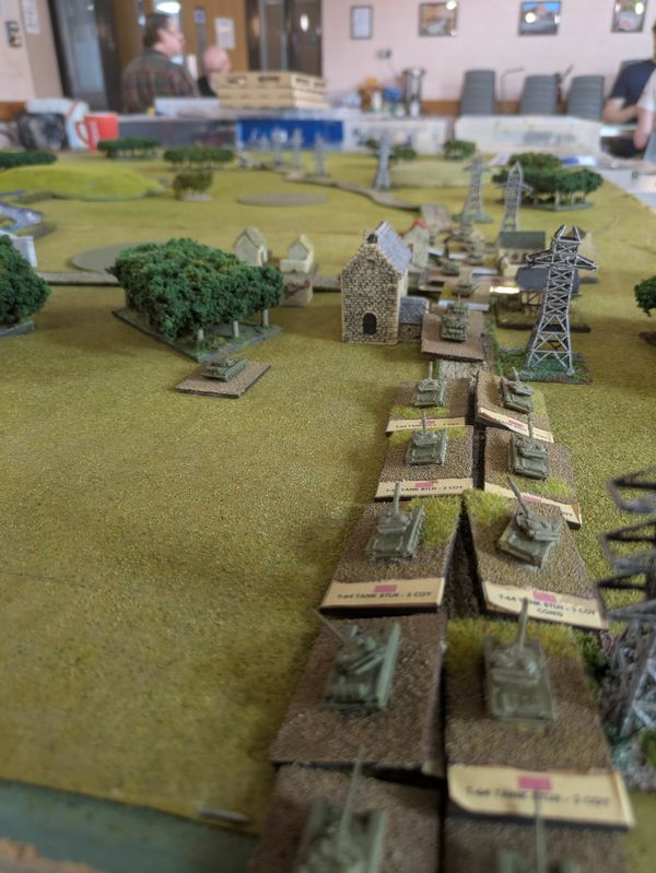 Close-up of a column of tanks advancing through terrain past a church and electricity pylons